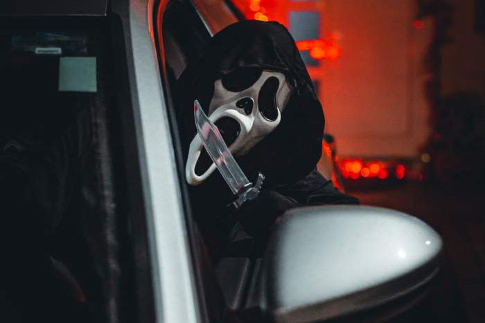 Ghostface sitting in a car and leaning out the windows with a knife in his hand.