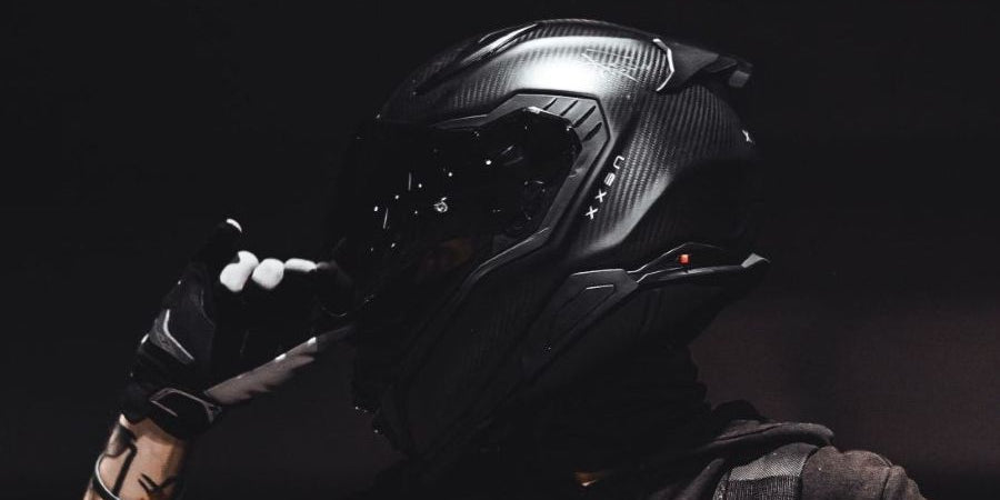 A person wearing a black full-face motorcycle helmet with a visor, black gloves, and a dark hoodie, posed against a dark background.