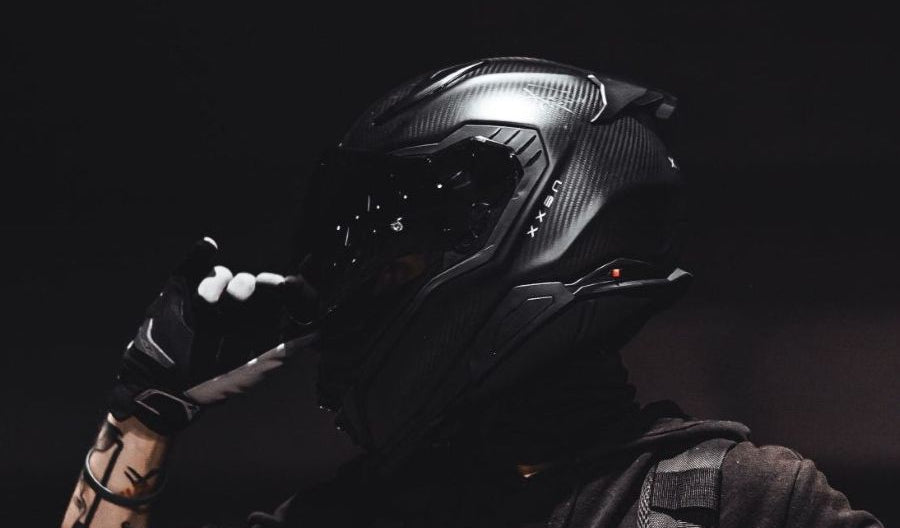A person wearing a black full-face motorcycle helmet with a visor, black gloves, and a dark hoodie, posed against a dark background.