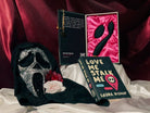 The Callahan silicone toy vibrator in the opened book-like package for Love Me Stalk Me by Laura Bishop and the gold bracelet next to a ghostface mask and the book Love Me Stalk Me.
