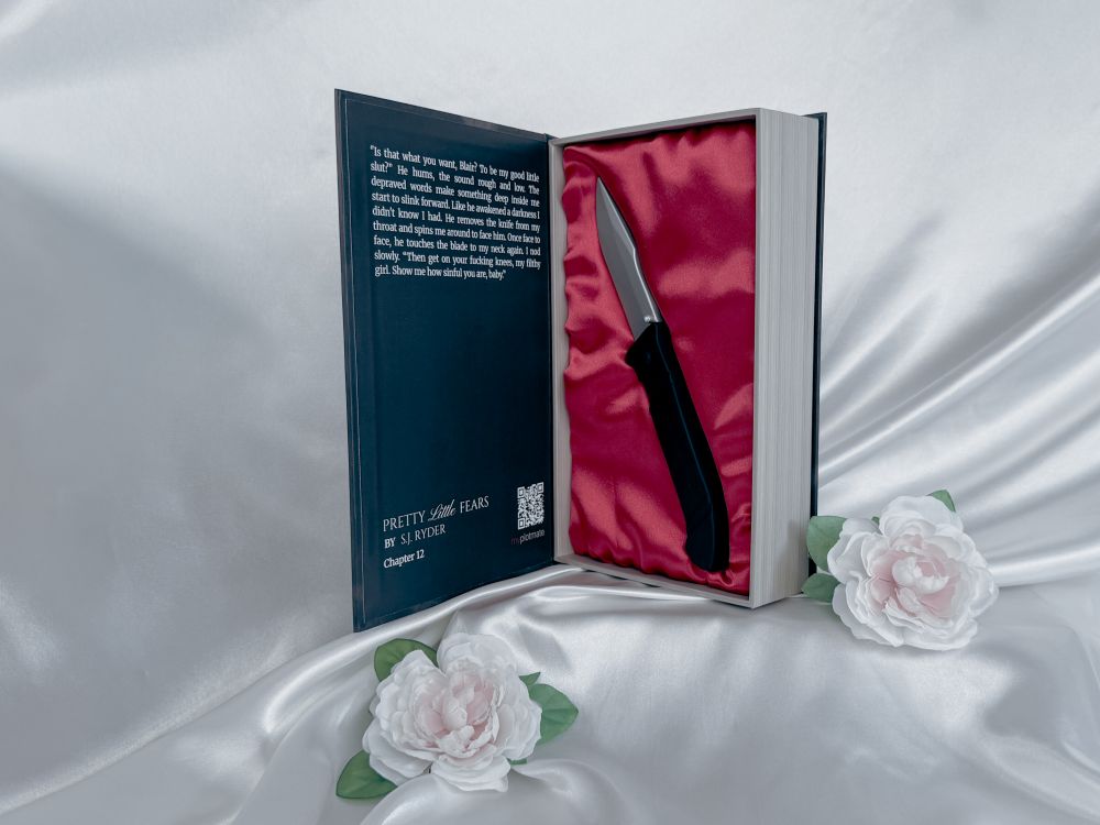 The silicone knife toy inside the opened book-like package for Pretty Little Fears by S.J. Ryder next to two white roses.