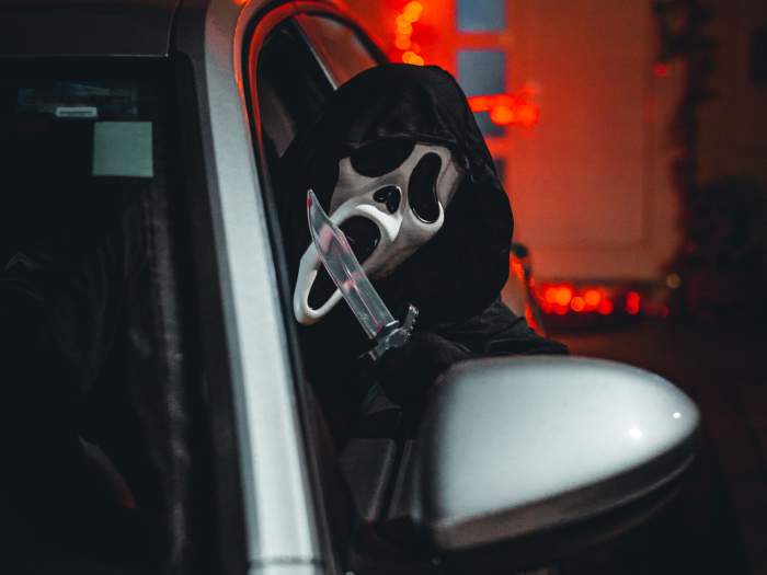 Ghostface sitting in a car and leaning out the windows with a knife in his hand.