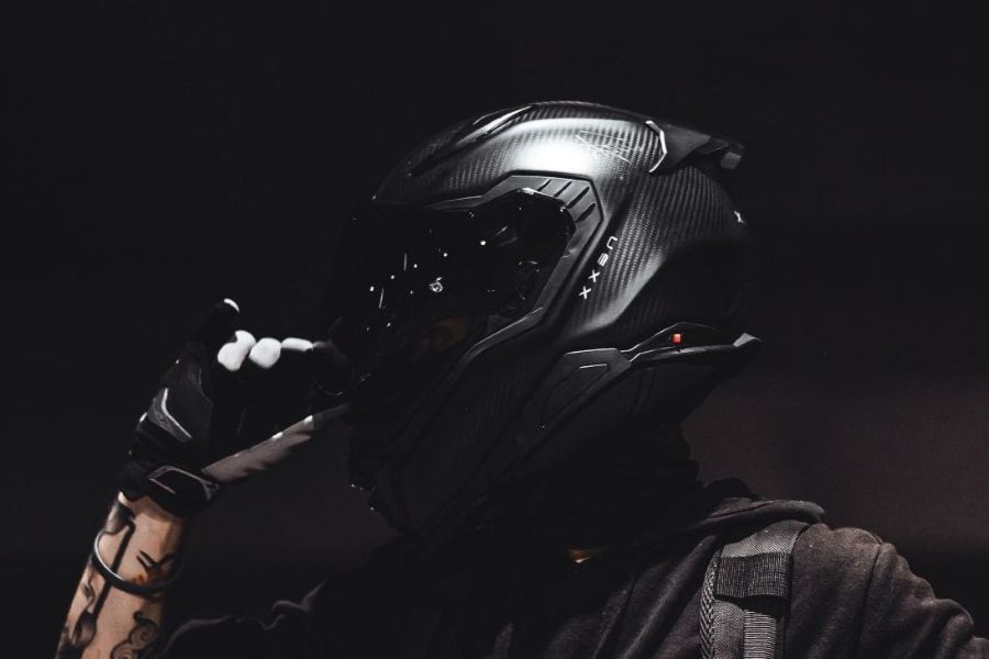 A person wearing a black full-face motorcycle helmet with a visor, black gloves, and a dark hoodie, posed against a dark background.