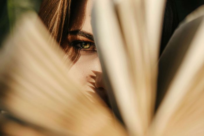 A close-up lifestyle shot of a woman's face partially obscured by the pages of an open dark romance book, with warm sunlight and soft focus.